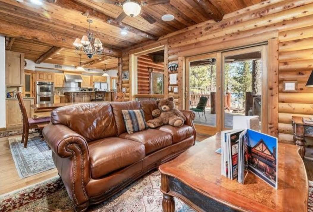 A log-walled living room with a plank ceiling, wood-cased doors and a kitchen of knotty wood cabinets beyond