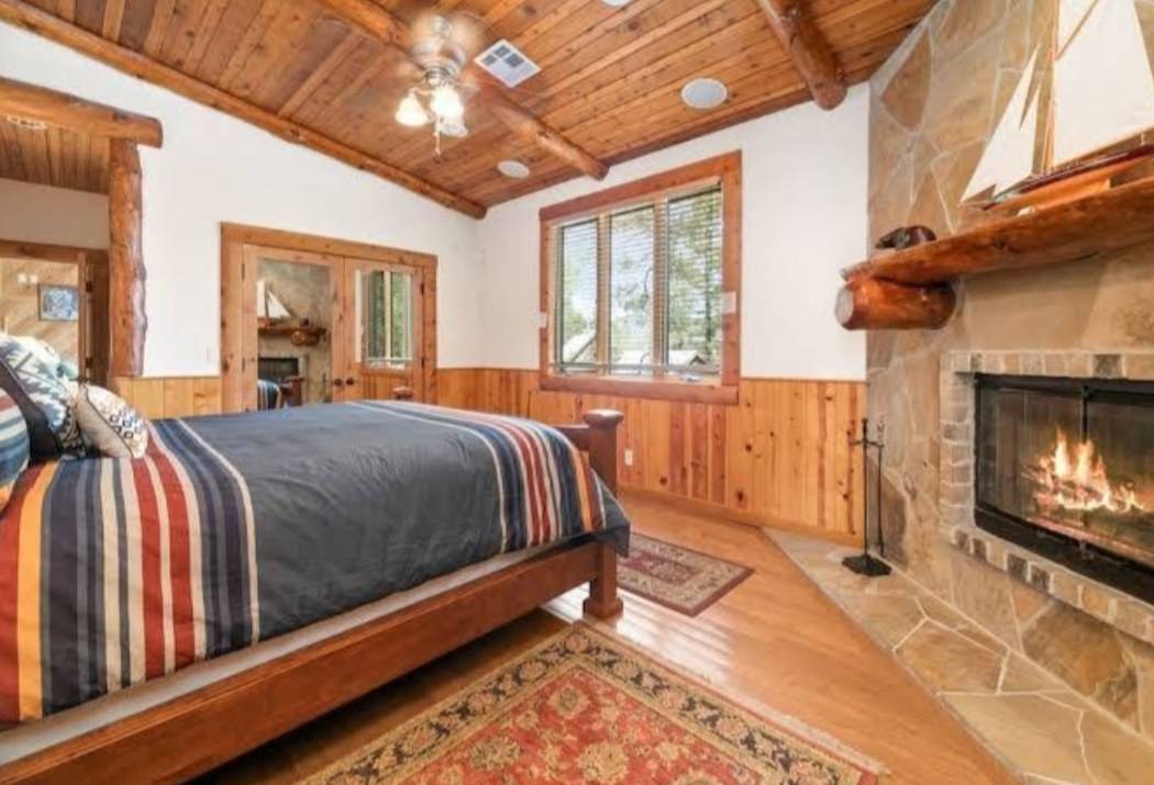 A bedroom with a log mantel over a stone fireplace, wood wainscoting and a plank ceiling