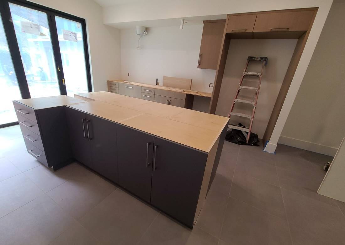 A dark flat-panel kitchen island and light oak wall cabinets, with plywood tops awaiting stone
