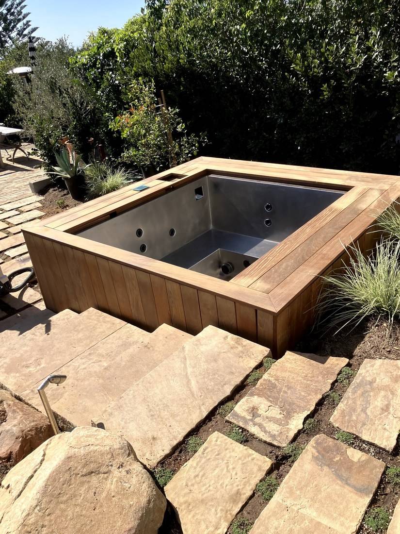 A square stainless steel spa clad in vertical hardwood boards with a wide mitered hardwood cap, beside stone steps