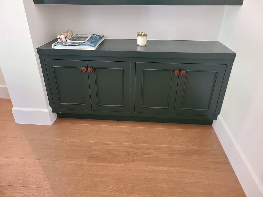 A low dark green painted cabinet with four paneled doors fitted into an alcove