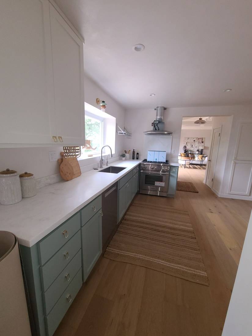 A long kitchen run of sage-green Shaker base cabinets with brass pulls under a white counter, and white wall cabinets