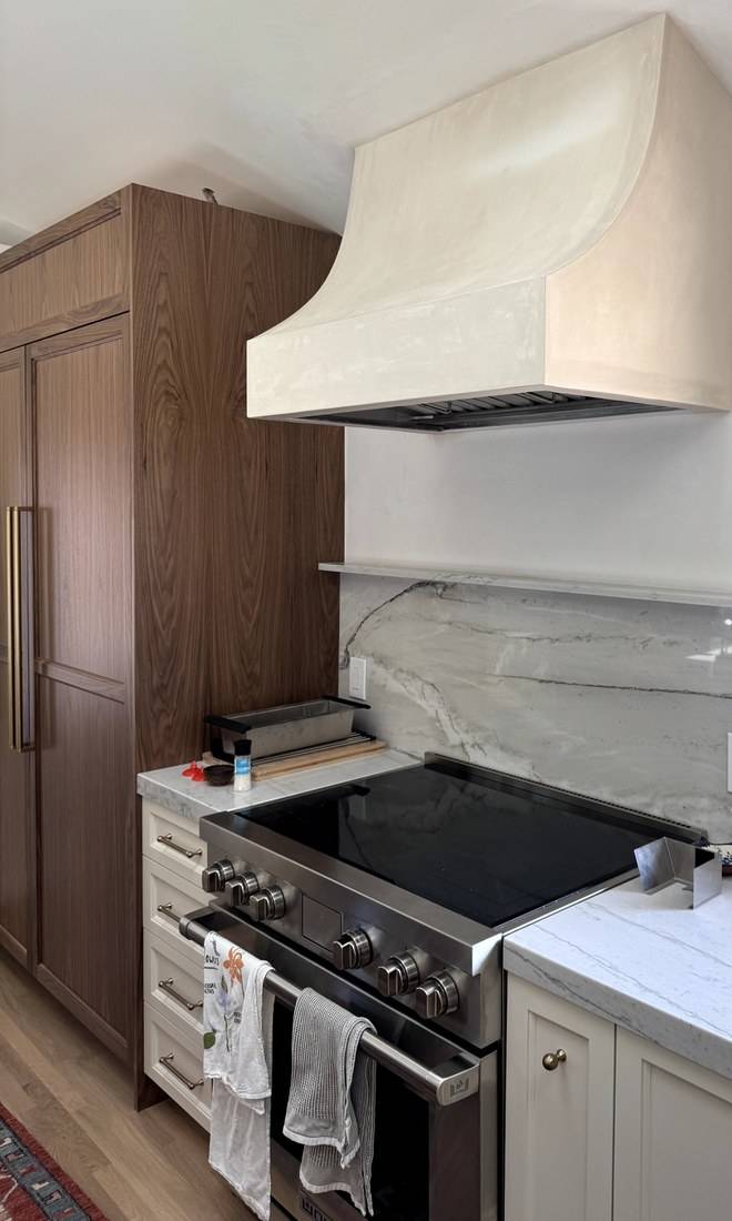 A tall walnut cabinet wall with frame-and-panel doors and brass pulls, beside a range under a plaster hood