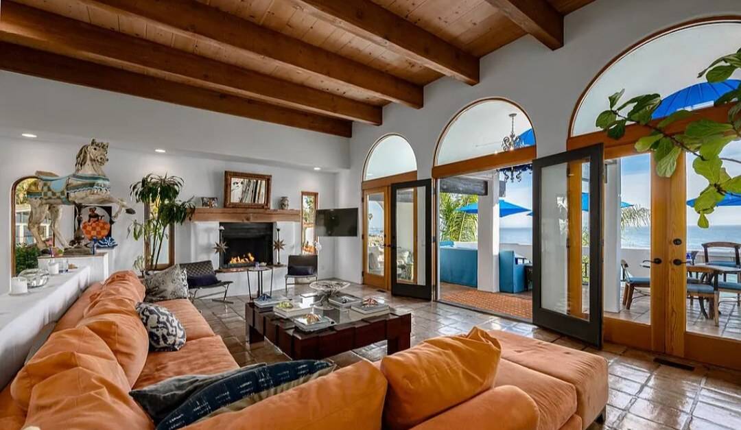 A living room with exposed wood beams, a plank ceiling, a wood mantel and arched wood-framed glass doors open to an ocean terrace
