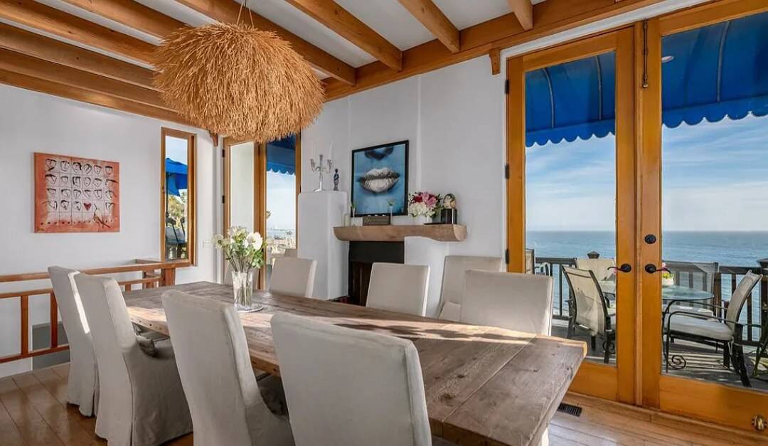 A dining room with exposed beams, a wood mantel shelf and wood-framed glass doors facing the ocean