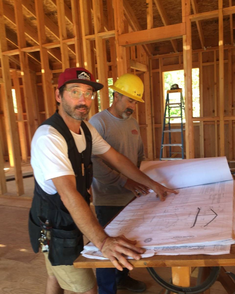 Matt Newman in a red cap and tool vest at a plan table inside a framed house, beside a man in a hard hat reading the drawings