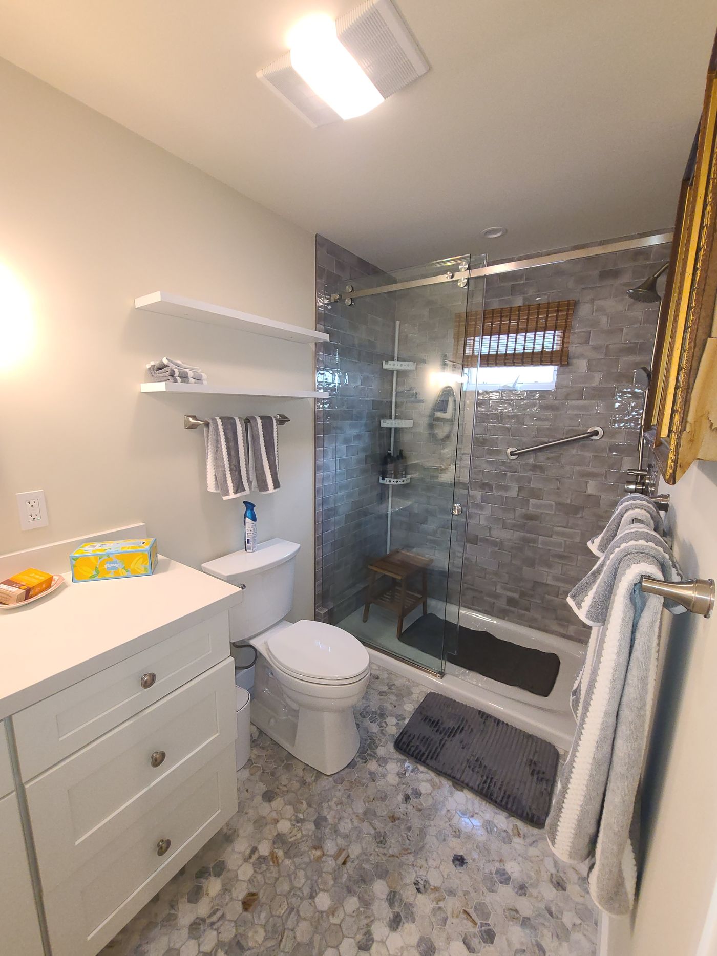 A bathroom with a walk-in shower in glazed gray subway tile behind a sliding glass door, a white Shaker vanity, floating white shelves and a gray marble hexagon mosaic floor