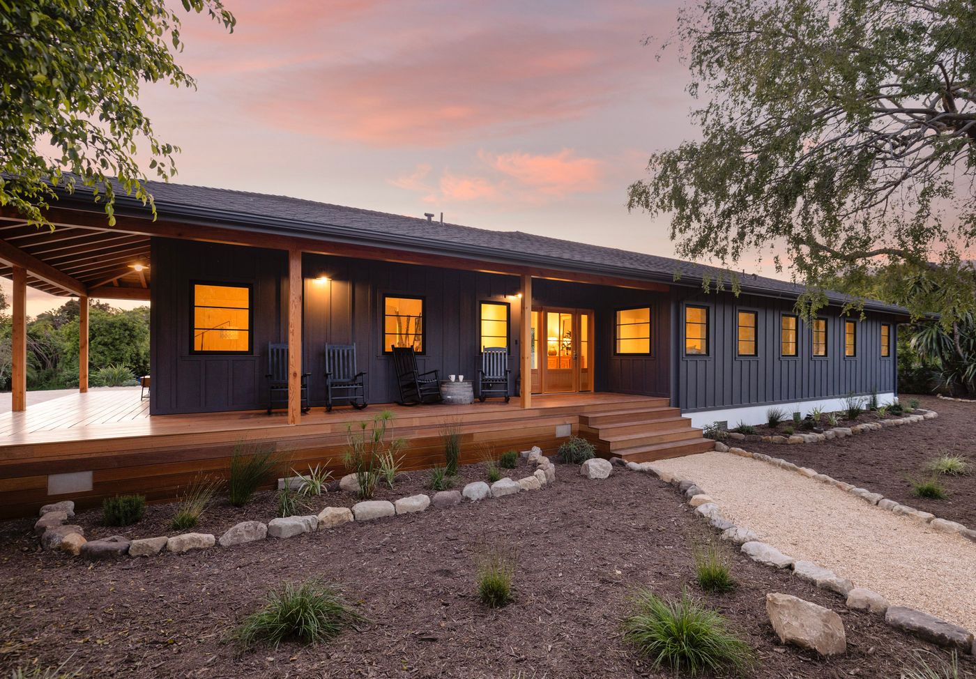 The front of the Loureyro Property at dusk: dark board-and-batten siding, black-framed windows glowing warm, and a long covered porch on timber posts above a gravel path edged in fieldstone