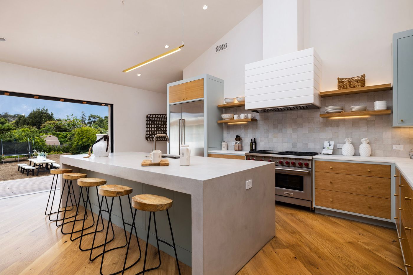 The kitchen: a concrete-finish waterfall island with oak stools, oak drawer fronts in gray-green frames, a white paneled range hood over handmade tile, and the glass wall slid open to the garden