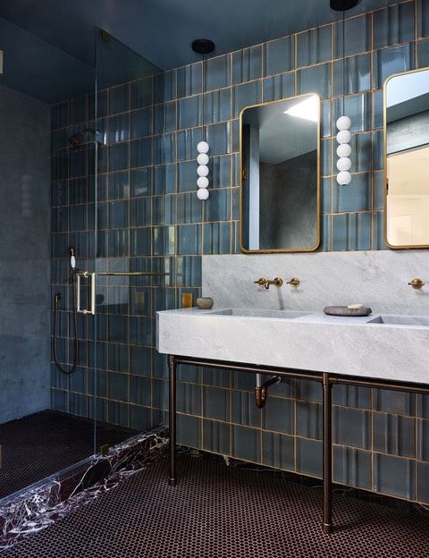 A bathroom lined in blue glazed tile with a stone trough sink and backsplash on a dark metal stand, brass wall-mounted faucets and mirrors, a glass shower with a veined dark stone curb, and a dark penny-round tile floor