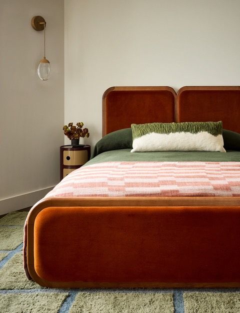 A bedroom with a rust velvet bed, a brass wall sconce and a green patterned rug