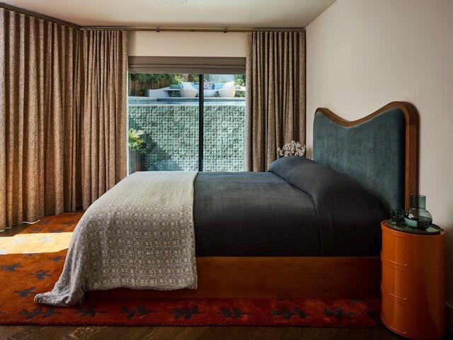 A bedroom with floor-to-ceiling curtains, a sliding glass door looking onto a green tiled garden wall, a teal upholstered bed and dark wood floors