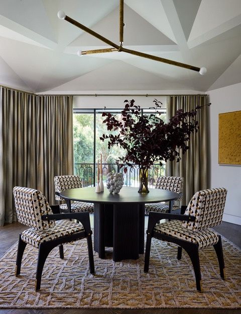A dining room with a faceted white ceiling, a brass pendant light, a round dark table with patterned chairs, and a tall window wall with a railed balcony beyond