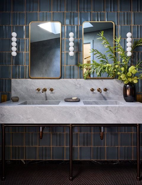 A double trough sink carved from gray stone with a matching backsplash, two sets of brass wall-mounted faucets, brass-framed mirrors and globe sconces on blue glazed tile