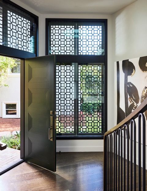 An entry hall with a tall dark paneled front door open to the garden, a full-height window with a geometric lattice screen, and a dark wood stair with a black metal rail