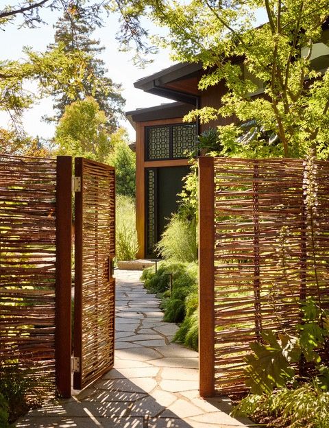 A garden gate of woven branches set in weathered steel frames, open onto a flagstone path through planting to a front door with a geometric window screen