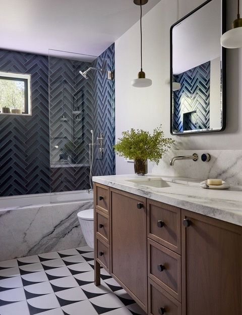 A bathroom with dark blue herringbone tile around a tub and shower, a veined white stone tub front and vanity top, a wood vanity with dark knobs, a wall-mounted faucet and a black and white patterned floor