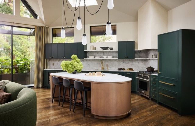 A kitchen with dark green cabinets and a paneled refrigerator, a veined stone backsplash with open shelves, a stainless range, and a rounded island with a white stone top and a copper-clad base, under pendant lights and a skylight