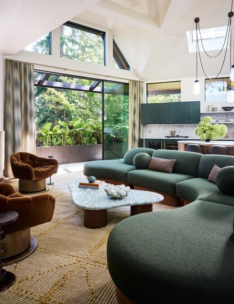 A living room with a large window wall to a planted terrace, a curved green sofa and a stone coffee table, opening to a kitchen with dark green cabinets, a stone backsplash and a skylight