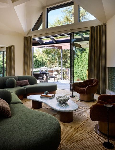 A living room with tall folding glass doors open to a covered terrace under a trellis, clerestory windows above, a curved green sofa and velvet swivel chairs