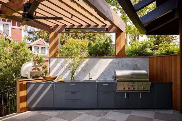 An outdoor kitchen under a wood trellis with dark base cabinets, a sink, a built-in grill, a pizza oven, a veined stone backsplash and wood-clad walls, on a stone-paved terrace