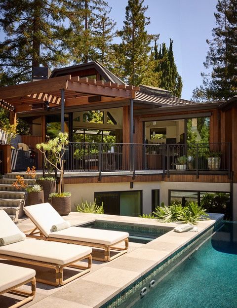 A stone pool deck with lounge chairs beside a pool and spa edged in green tile, below a wood-clad house with a railed upper terrace and a trellis