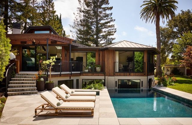 The back of a two-story house with vertical wood siding, a standing-seam metal hip roof and a wood trellis over a railed terrace, with stone steps down to a stone-paved pool deck and a rectangular pool