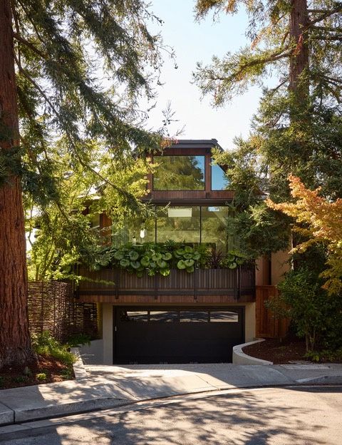 The street front of a house among tall redwoods: a dark garage door below a wood-clad upper floor with large windows and a planted balcony