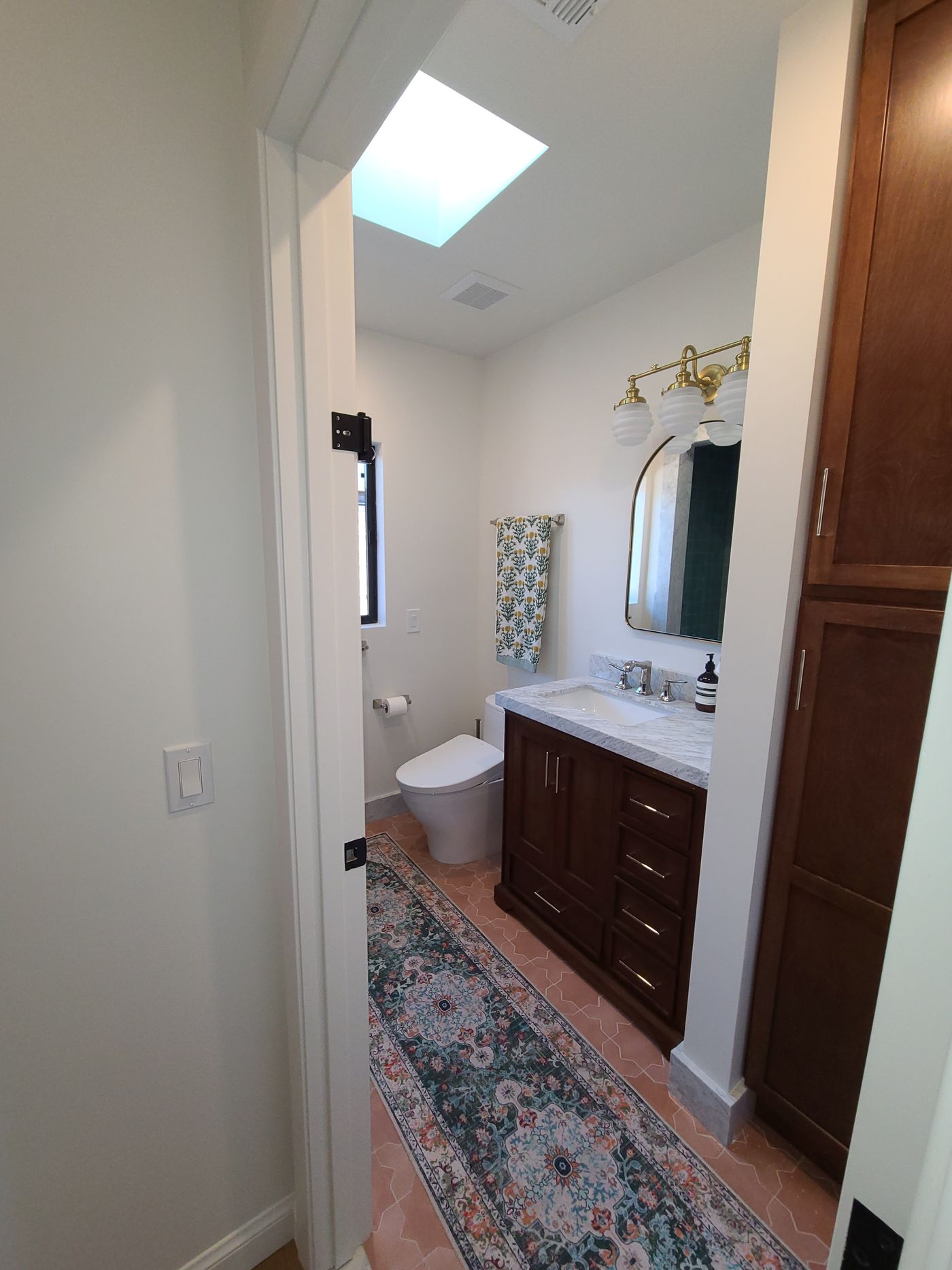 A bathroom under a skylight with a terracotta tile floor, a dark walnut vanity with a marble top, an arched brass mirror and brass light fixture, and a tall walnut linen cabinet
