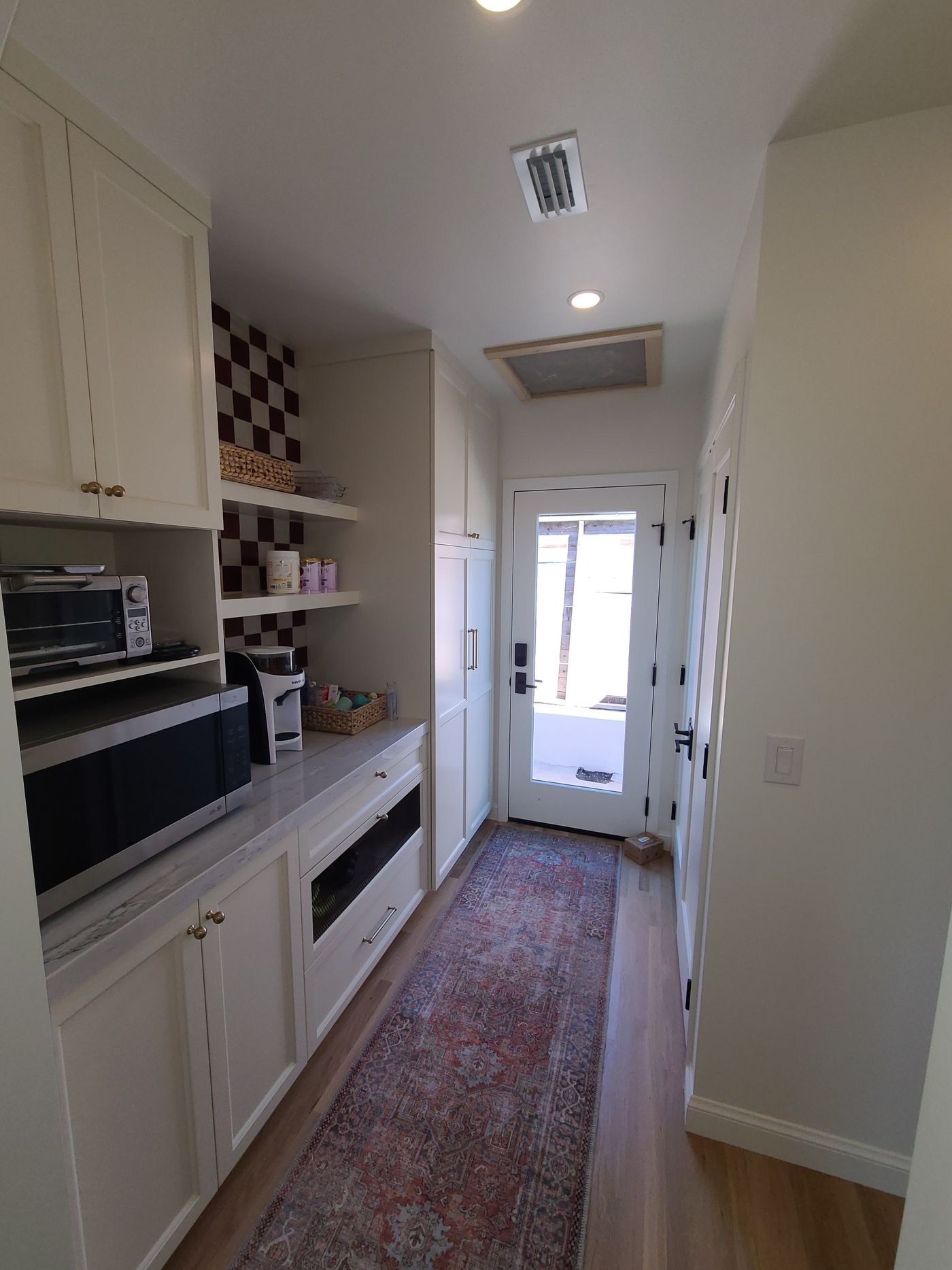 A narrow butler's pantry with cream Shaker cabinets, open shelves on a checkerboard tile wall, a stone counter, and a glass door at the end of a runner-lined hall