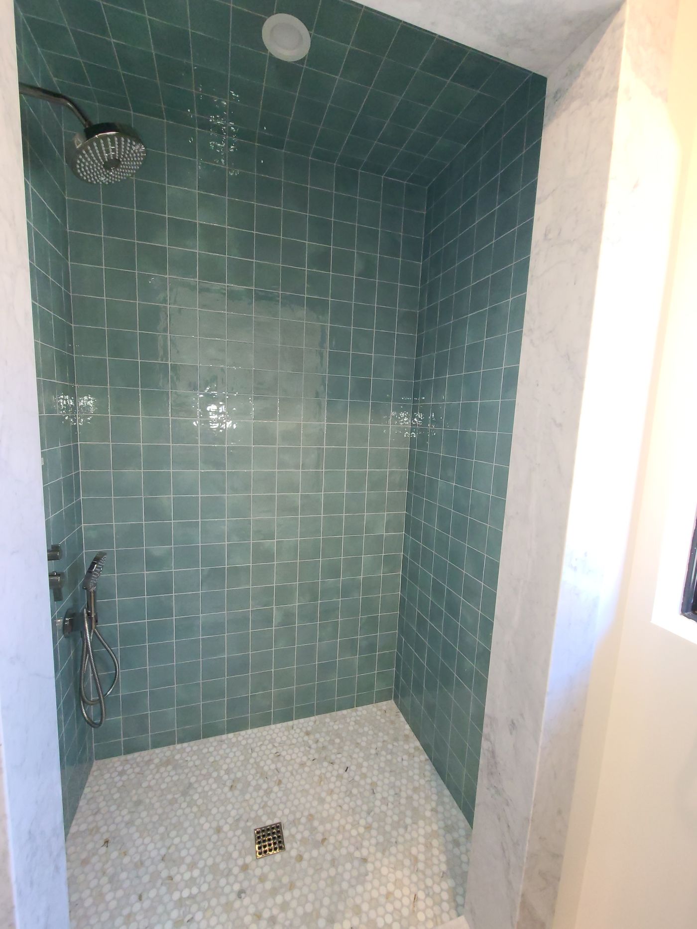 A walk-in shower lined floor to ceiling in glazed green square tile, framed in white marble, with a penny-round mosaic floor and a rain head