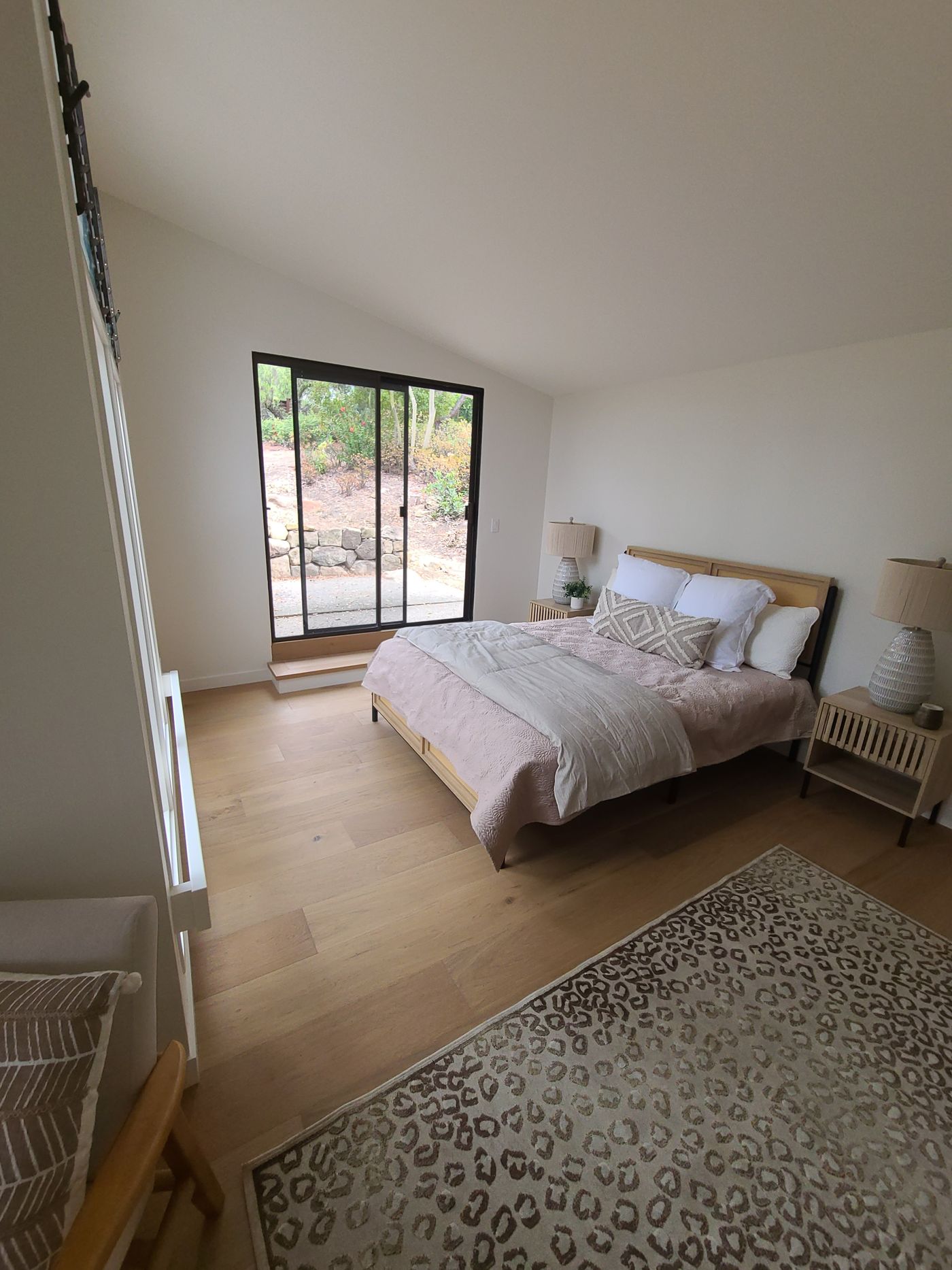 A bedroom with a vaulted ceiling, wide-plank wood floors, and a black-framed sliding glass door above a wood step, looking out to a garden and a stone wall