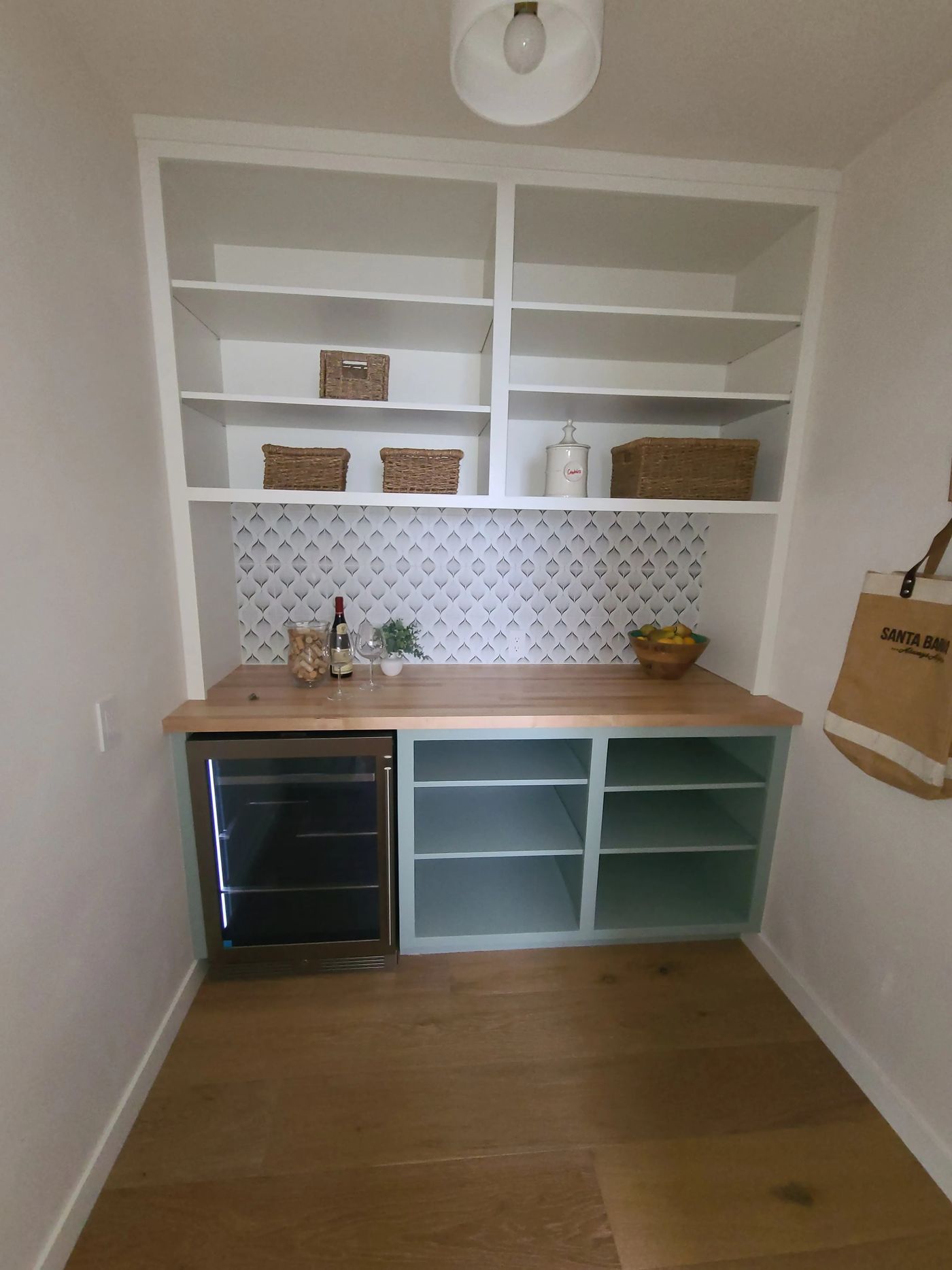 A walk-in pantry with white open shelving above a patterned tile backsplash, a butcher-block counter, sage-green open base cabinets and a stainless wine refrigerator
