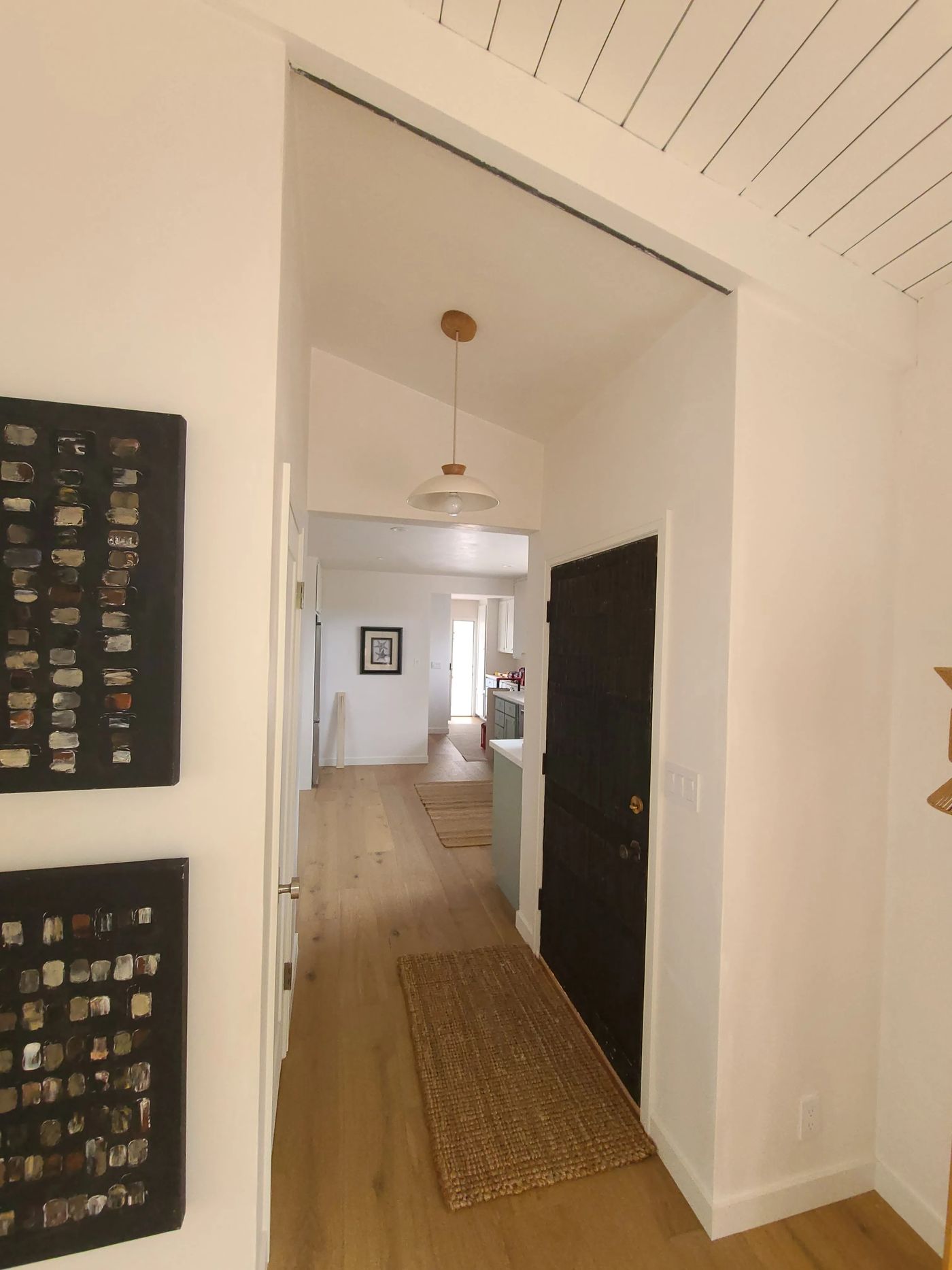 An entry hall with a dark wood front door, a pendant light under a vaulted ceiling, white walls and wide-plank wood floors running through to the kitchen