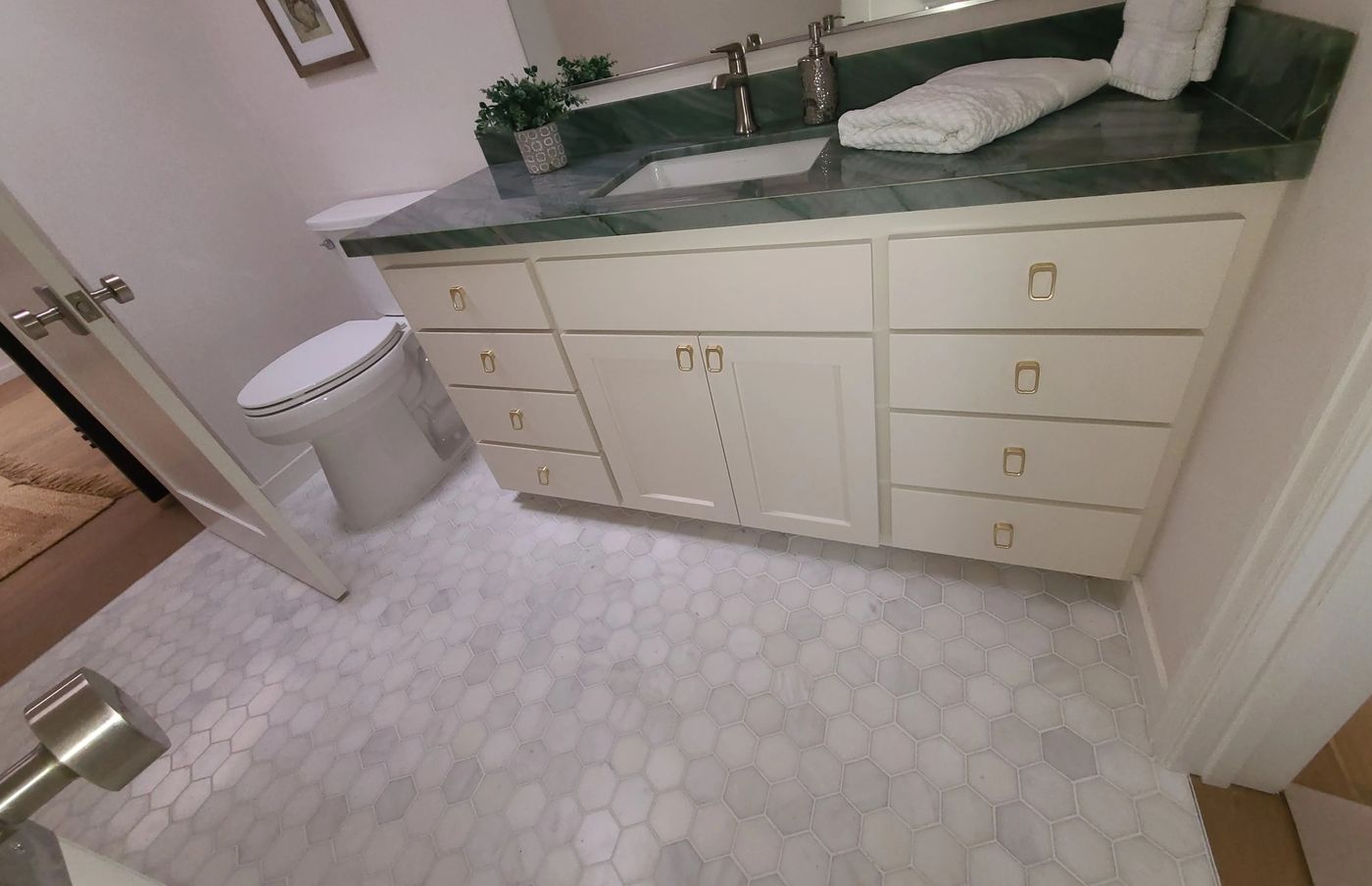 A bathroom vanity with a polished green veined stone top and undermount sink, cream Shaker drawers and doors with brass pulls, beside a toilet, on a white and gray marble hexagon mosaic floor