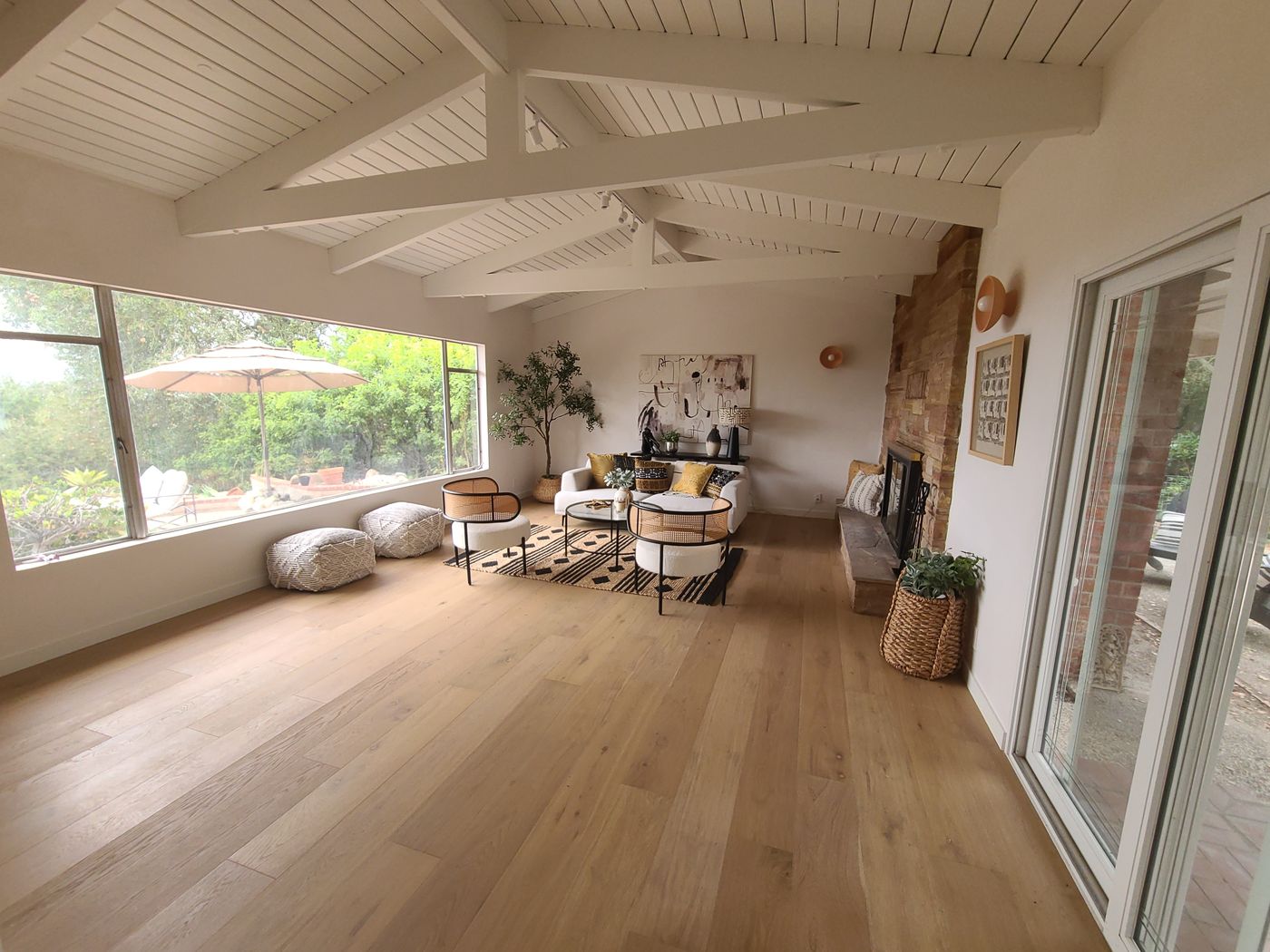 A living room with a vaulted, white-painted board ceiling and exposed trusses, wide-plank wood floors, a long picture window to the garden, a stacked stone fireplace, and sliding glass doors