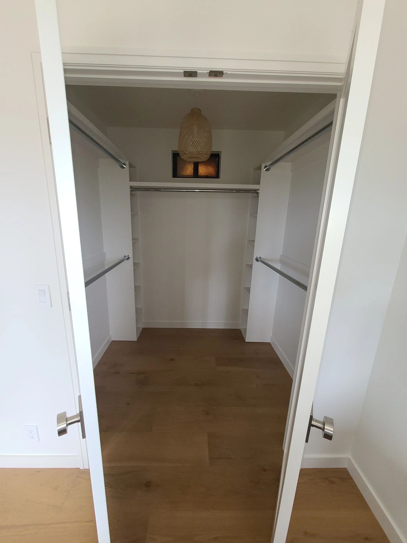 A walk-in closet seen through a pair of white doors, with white built-in shelf towers, double hanging rods on both sides and a woven pendant light